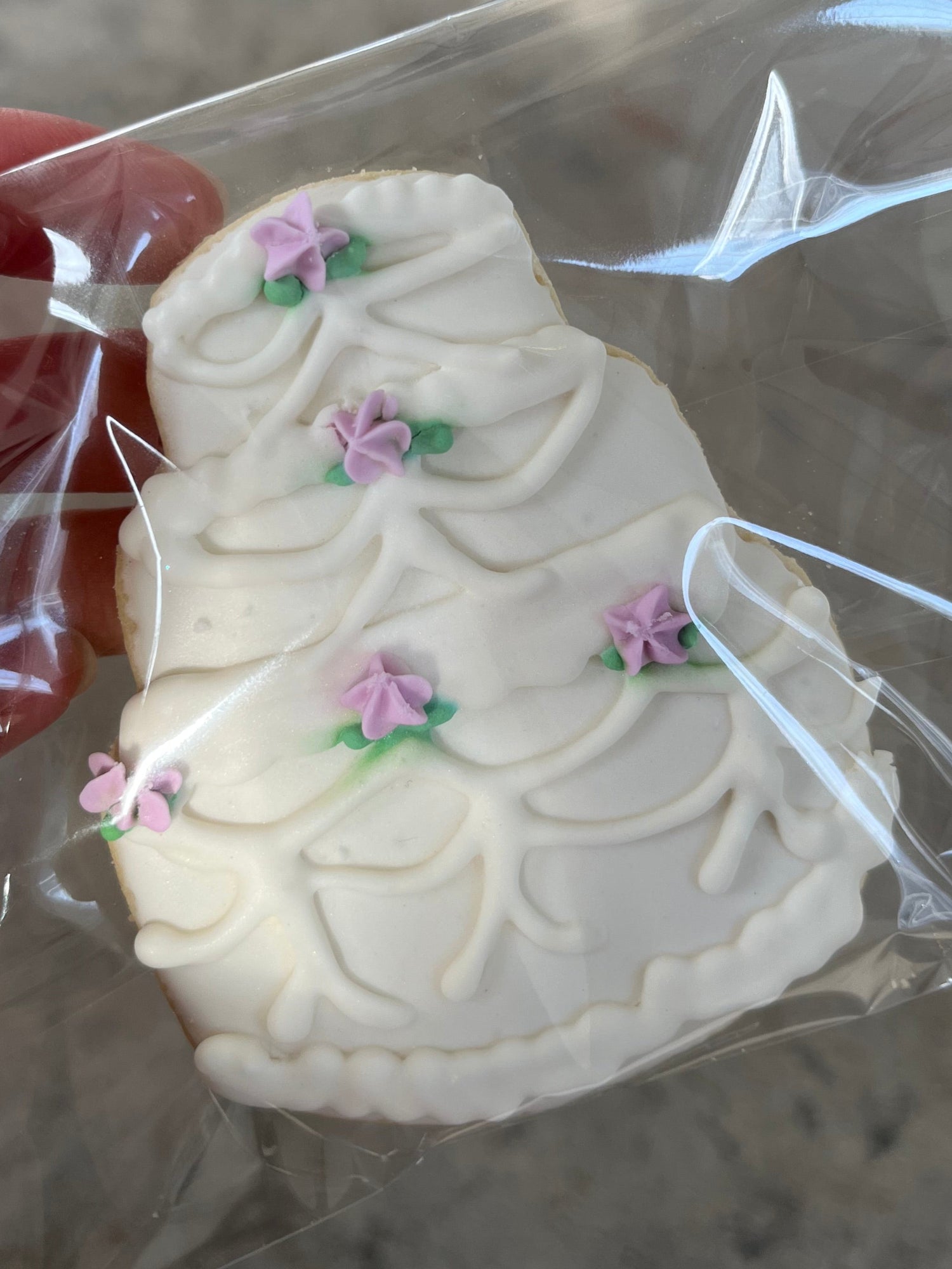 Wedding cake cookie with white icing and pink flowers in clear packaging held by a hand against a neutral background